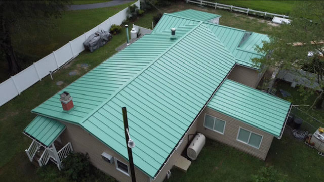 Aerial view of a single-story house with a light green metal roof, tan siding, and several attached sections, surrounded by white fencing and greenery in Talbot County, MD—perfect inspiration for your next roofing contractor project.