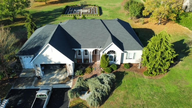 Aerial view of a single-story house in MD with a dark gray roof by a trusted roofing contractor Talbot County, attached garage, driveway with parked vehicle, landscaped yard, and a decorative garden featuring a maze pattern in the background.