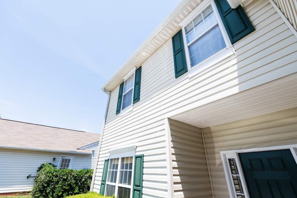 Close-up view of a beige two-story house with green window shutters and a green front door in Talbot County, MD, seen from a low angle on a sunny day. A nearby building hints at the expertise of a local roofing contractor.