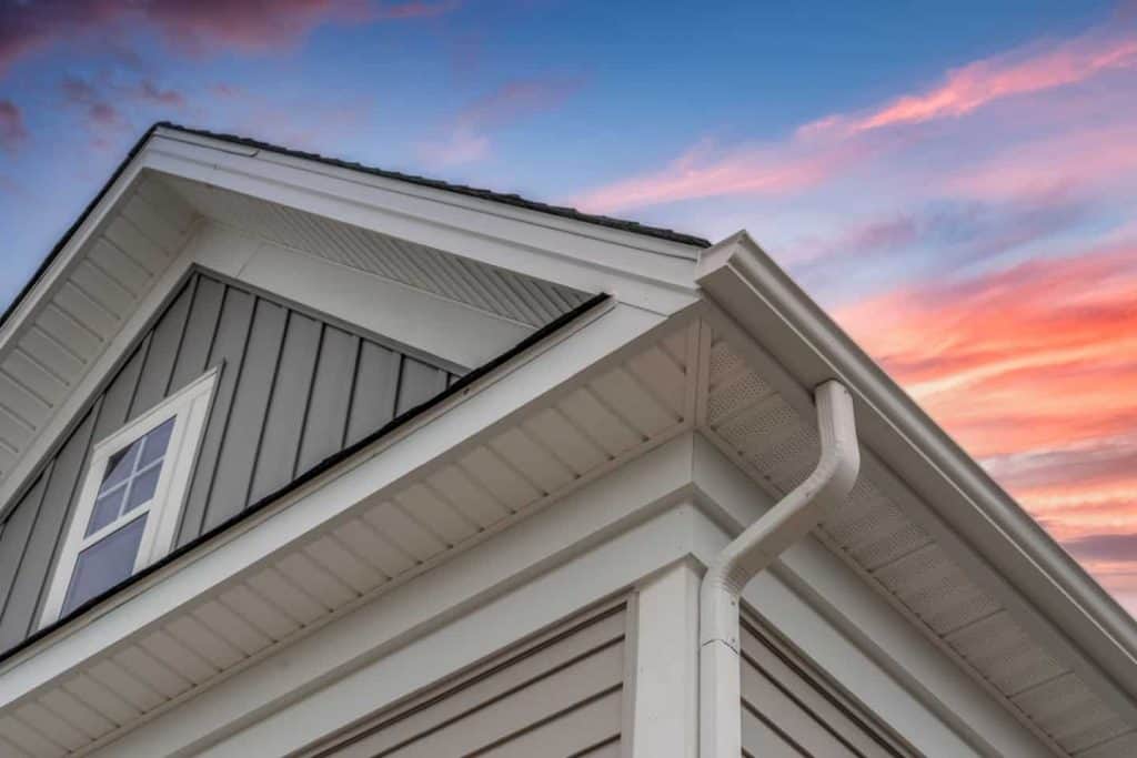 Close-up of a house’s roof and gutter system at sunset, featuring white trim and gray siding against a colorful sky. A trusted roofing contractor Talbot County, MD can help maintain this stunning look.