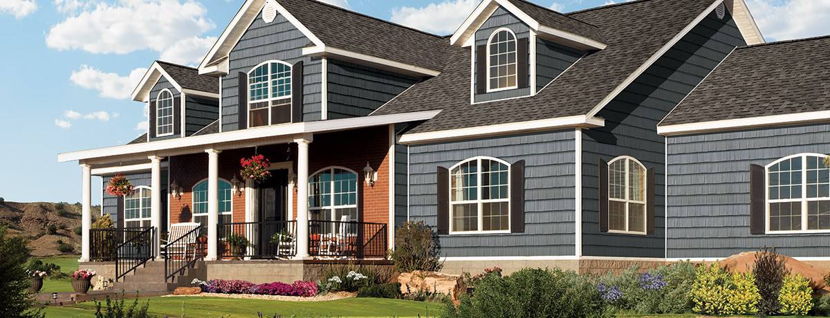 A large, two-story house with blue siding, white trim, and brick accents features dormer windows, a covered front porch with white columns, and a landscaped yard—perfect for any MD homeowner seeking a trusted roofing contractor in Talbot County.