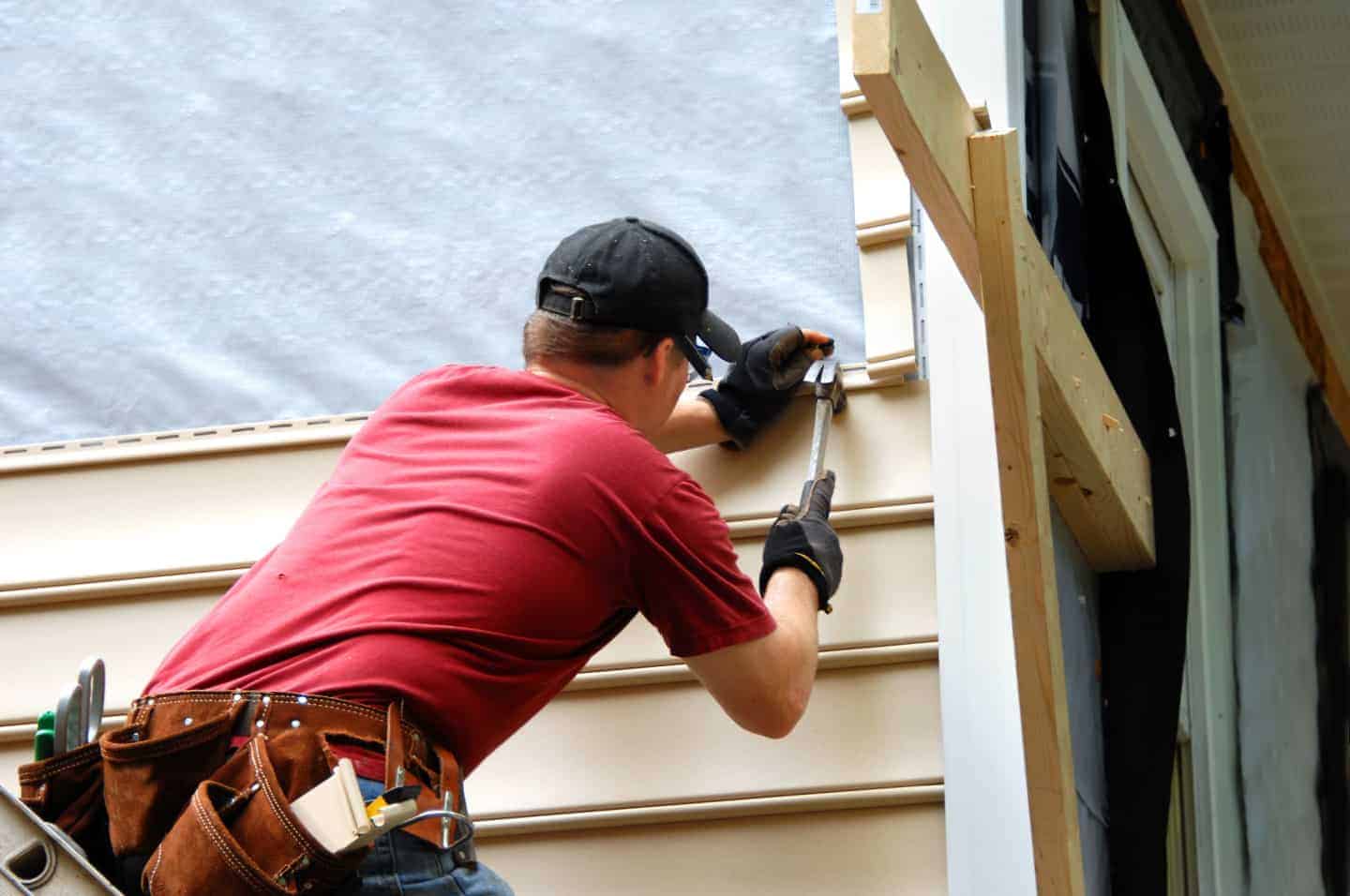 A person wearing a red shirt, black cap, and tool belt installs beige vinyl siding on a house, using a hammer and nails—much like a skilled roofing contractor in Talbot County, MD.
