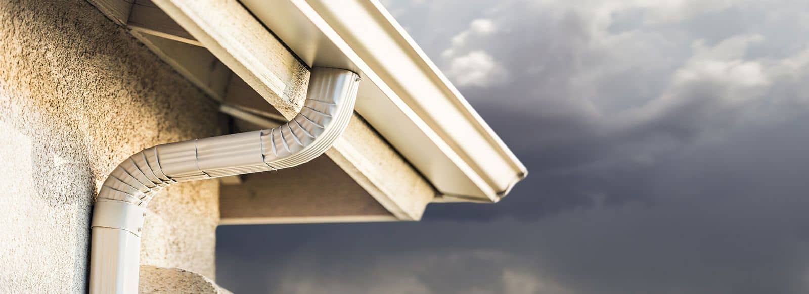 A close-up view of a house’s white rain gutter and downspout against a cloudy, dark sky, suggesting an approaching storm—an ideal reminder to call a roofing contractor Talbot County, MD for inspection. The building exterior appears textured and light-colored.