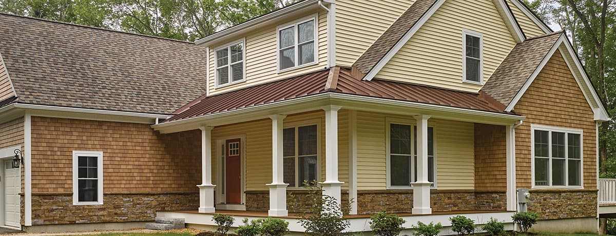 A beige and brown two-story house in MD with a covered front porch, white pillars, stone accents, and wood siding—showcasing quality by a trusted roofing contractor Talbot County—nestled among lush greenery and trees.