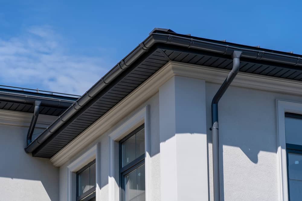 Close-up of a modern house corner featuring black rain gutters and downspouts, white walls, and large windows under a blue sky—quality work from a trusted roofing contractor in Talbot County, MD.
