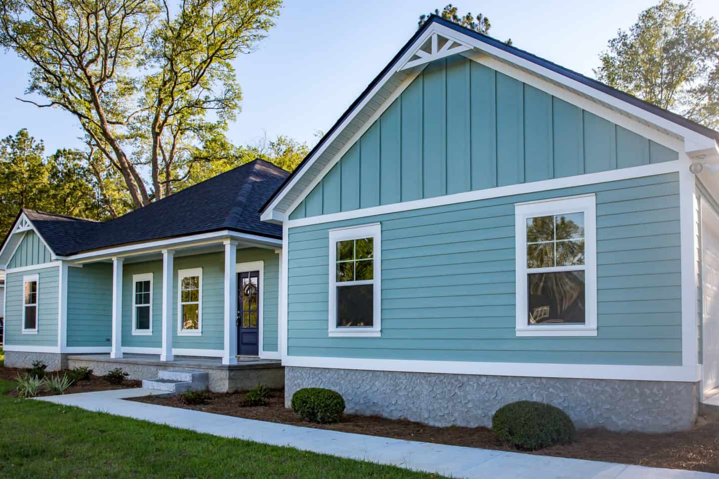 A single-story house in MD with blue-green siding, white trim, multiple windows, a dark roof installed by a trusted roofing contractor Talbot County. The yard features green grass, shrubs, and a concrete walkway leading to a small covered front porch.