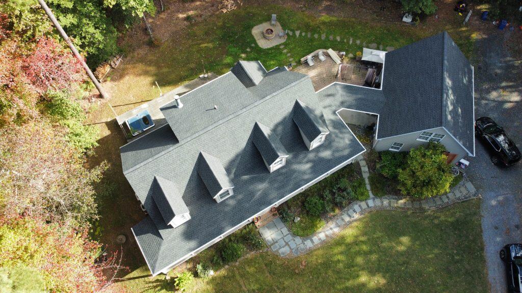 Aerial view of a house with multiple dormer windows, a connected garage, stone pathway, lush lawn, trees, and a backyard fire pit area—expertly maintained by a top roofing contractor in Talbot County, MD.