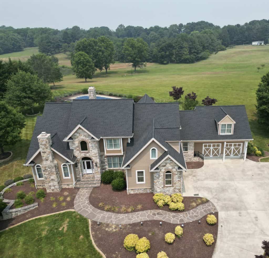 Aerial view of a large, two-story house with stone and beige siding, landscaped yard, curved walkway, attached garage, and new roof by a trusted roofing contractor Talbot County, MD—all surrounded by green fields and trees.