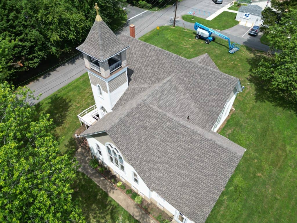 Aerial view of a white church with a tall steeple and gray shingle roof, surrounded by green grass and trees in Talbot County, MD—a beautiful landmark that could benefit from the expertise of a roofing contractor Talbot County, MD.