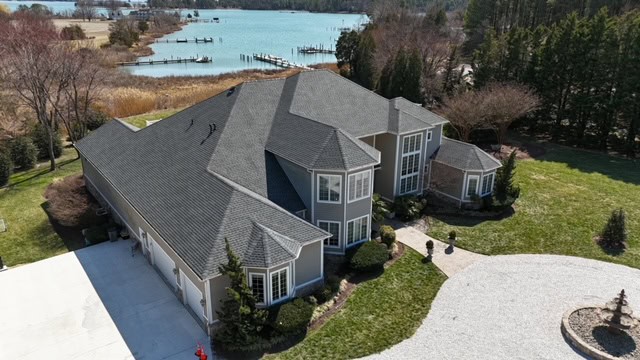 Aerial view of a large two-story house with gray roofing by a trusted roofing contractor Talbot County, MD, beige walls, landscaped lawns and trees, a circular gravel driveway, and a body of water in the background.