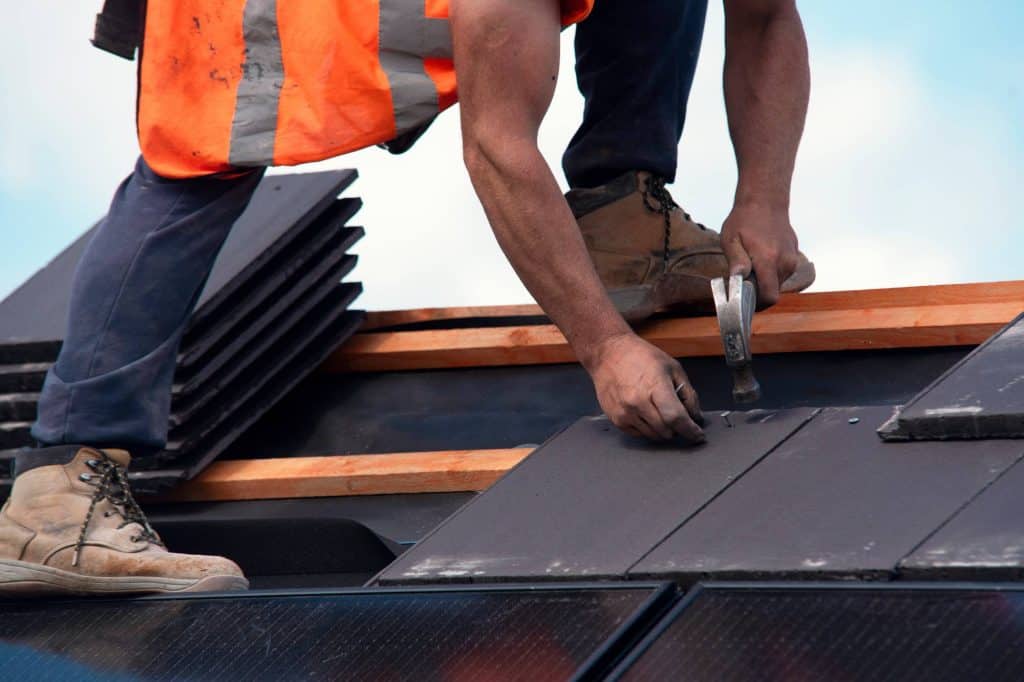A roofing contractor in Talbot County, MD, wearing an orange safety vest and boots, installs roof tiles with a hammer, securing them in place on a rooftop under a blue sky.
