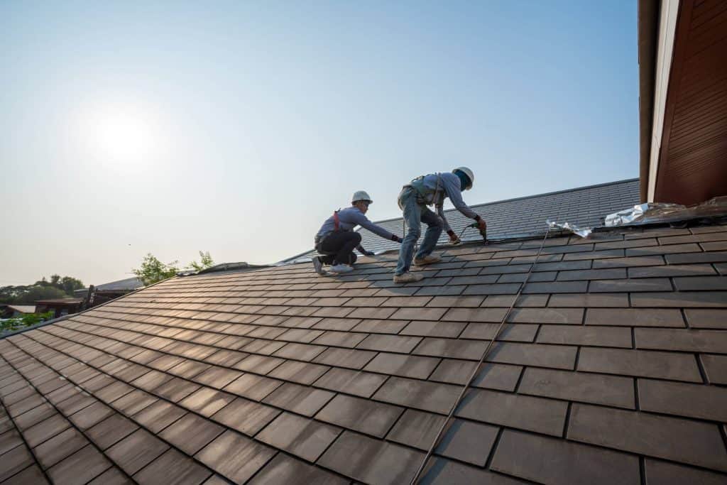 Two workers wearing safety harnesses and helmets install or repair shingles on a sloped roof under a clear sky, with sunlight in the background.