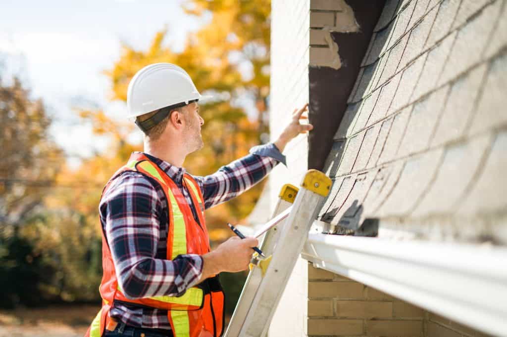 A construction worker in a hard hat and safety vest stands on a ladder, inspecting the roof and exterior wall of a house on a sunny day with autumn trees in the background.