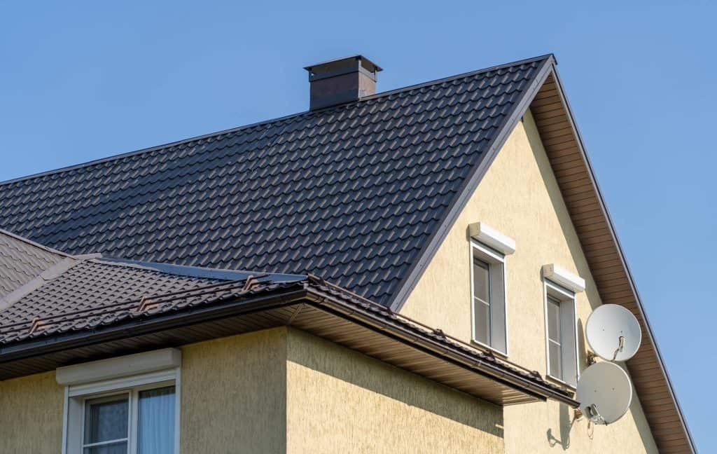 The upper part of a yellow house with a dark metal roof, white-trimmed windows, two satellite dishes, a chimney, and a clear blue sky in the background.