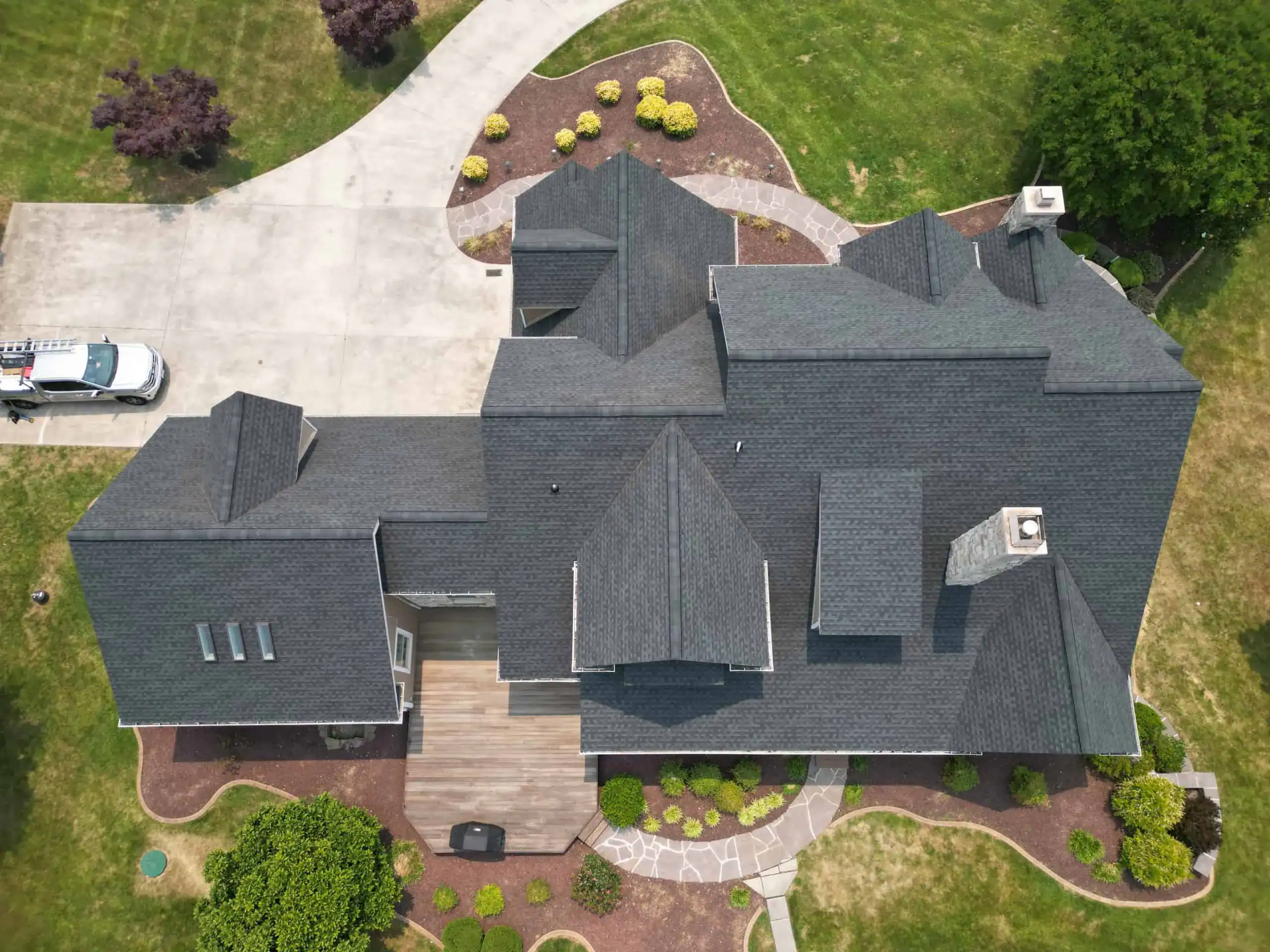 Aerial view of a large house with a dark gray roof, surrounded by green lawn, landscaped bushes, winding stone paths, and a circular driveway with a parked white vehicle.
