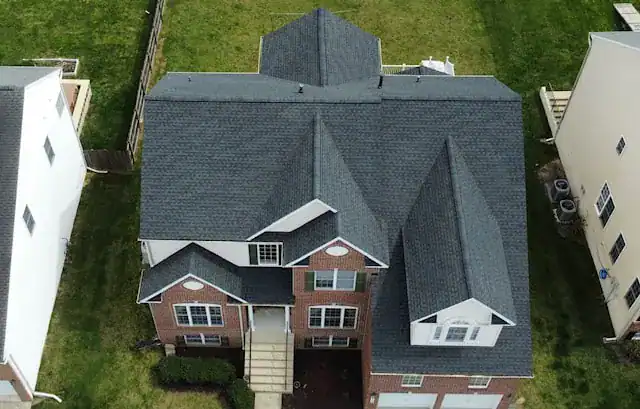 Aerial view of a two-story brick house with dark gray shingles, white trim, and an attached garage, surrounded by green grass and neighboring houses.
