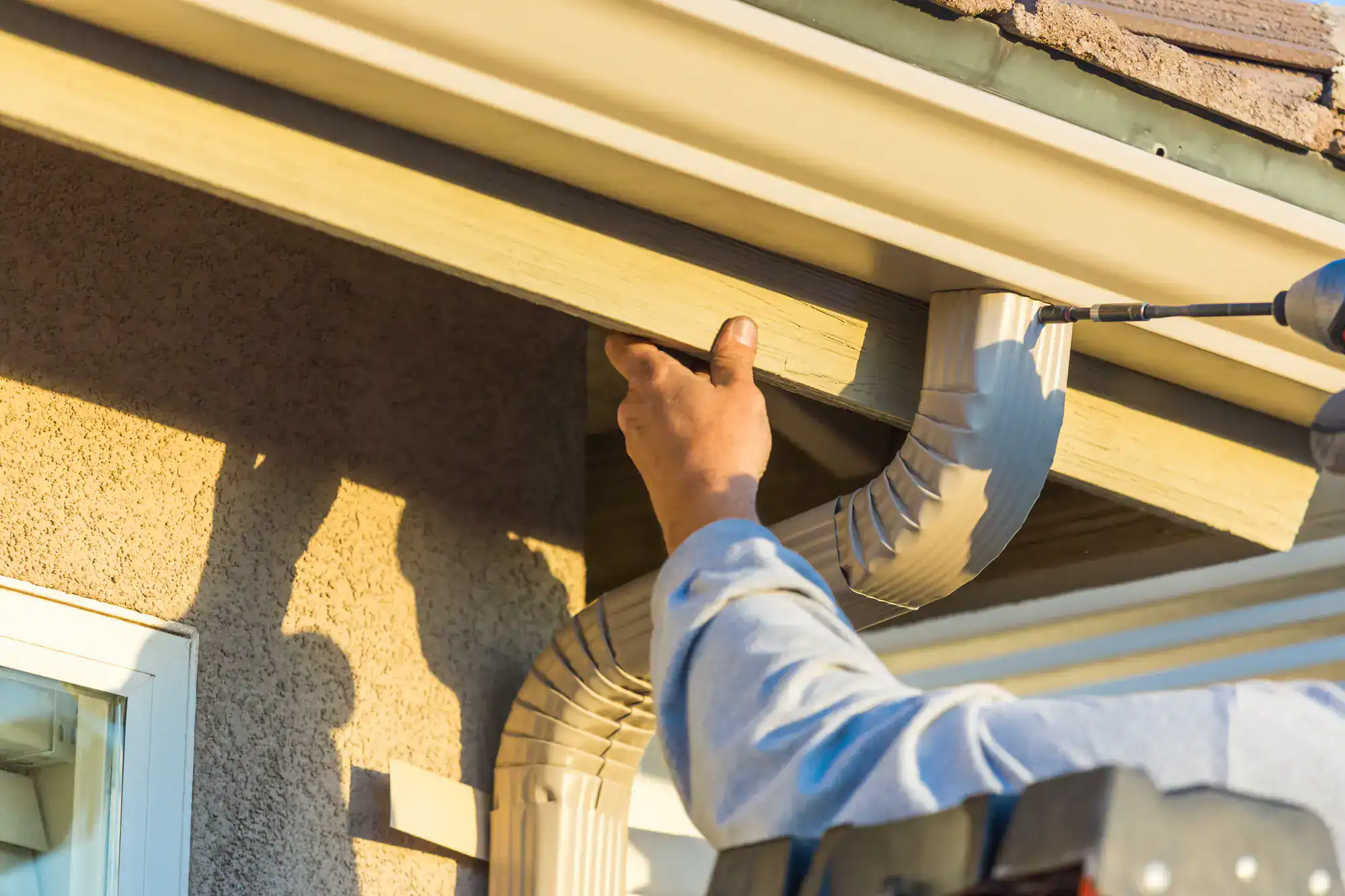 A worker on a ladder installs or repairs a beige rain gutter and downspout on the edge of a house roof, using a power drill. Sunlight casts shadows on the wall and highlights the worker’s hand.