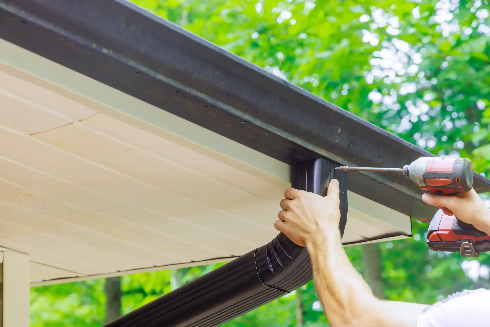 A person uses a power drill to attach a black downspout to the gutter system of a house, with green trees blurred in the background.