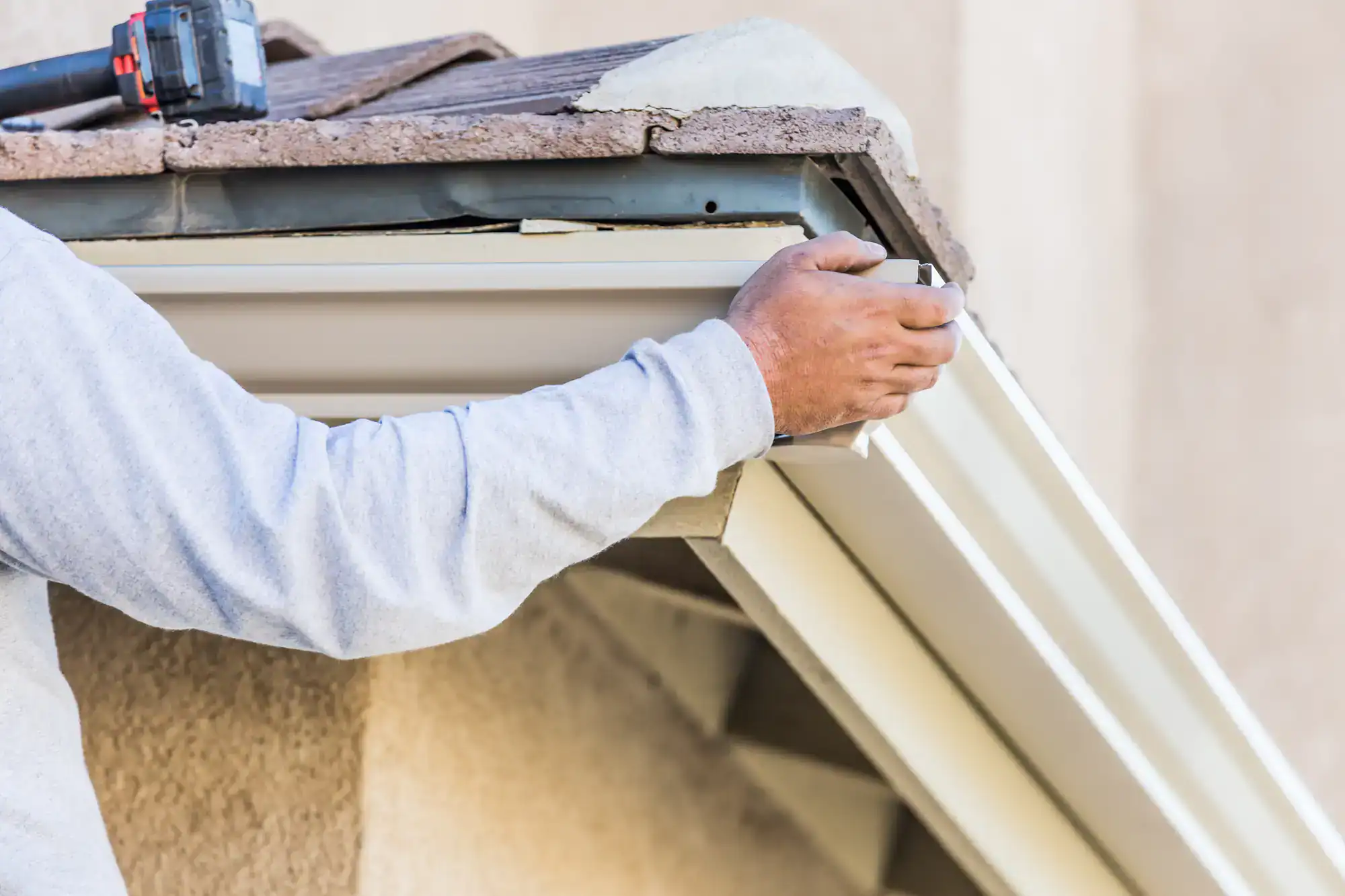 A person installing or repairing a white rain gutter on the edge of a roof, holding the gutter in place with one hand while using a tool. The person's face is not visible.
