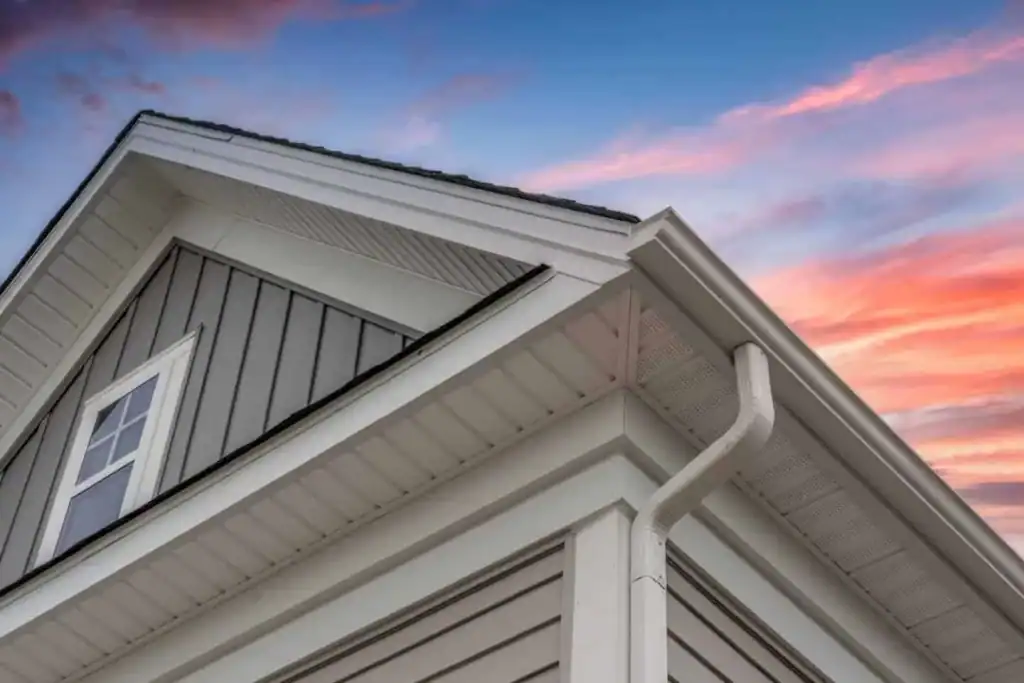 Close-up view of a house’s corner showing white gutters and gray siding, with a window and angled roofline, set against a colorful sunset sky with pink, orange, and blue hues.