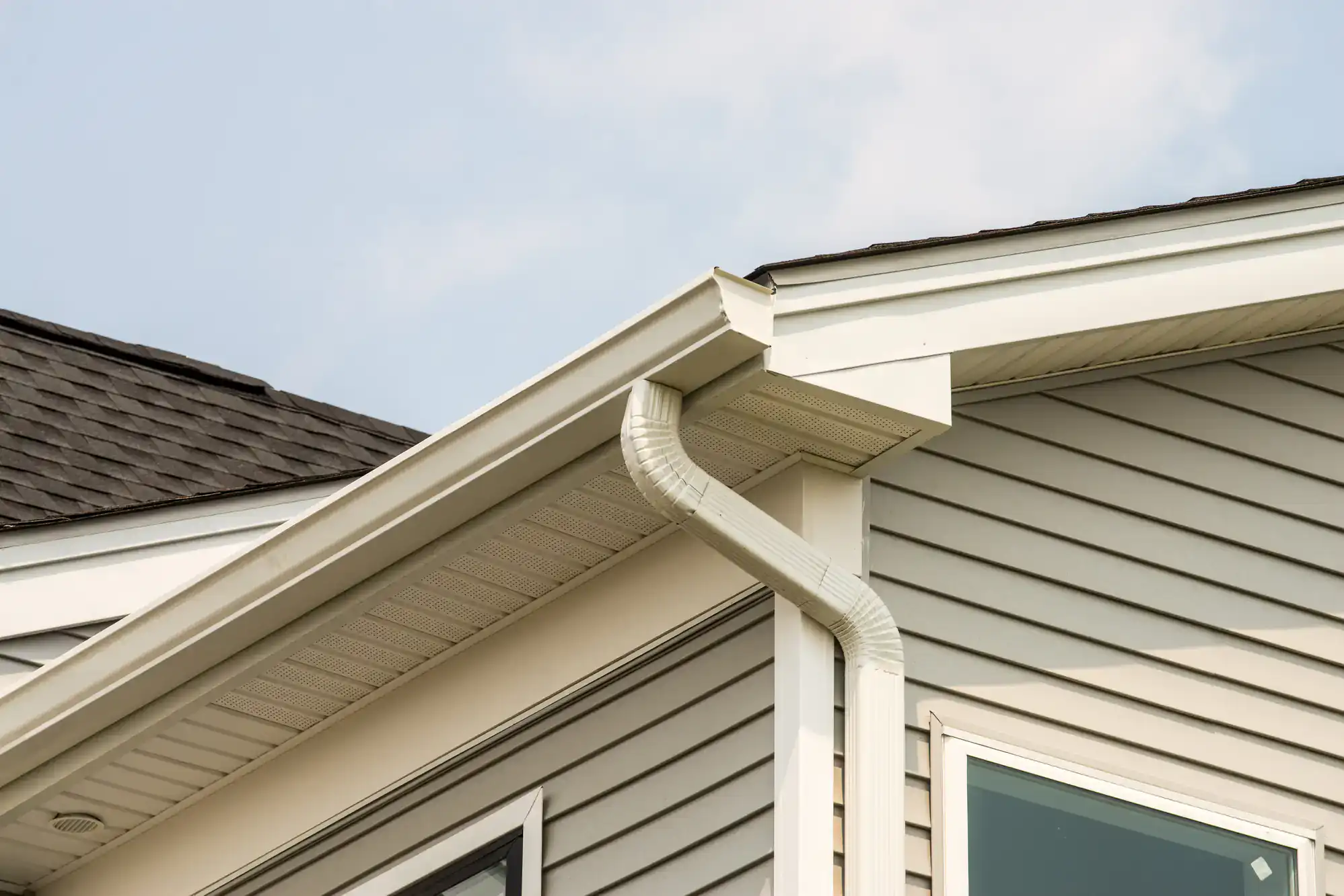 Close-up of a house corner showing gray vinyl siding, white gutter system, downspout, and black shingled roof against a partly cloudy sky.
