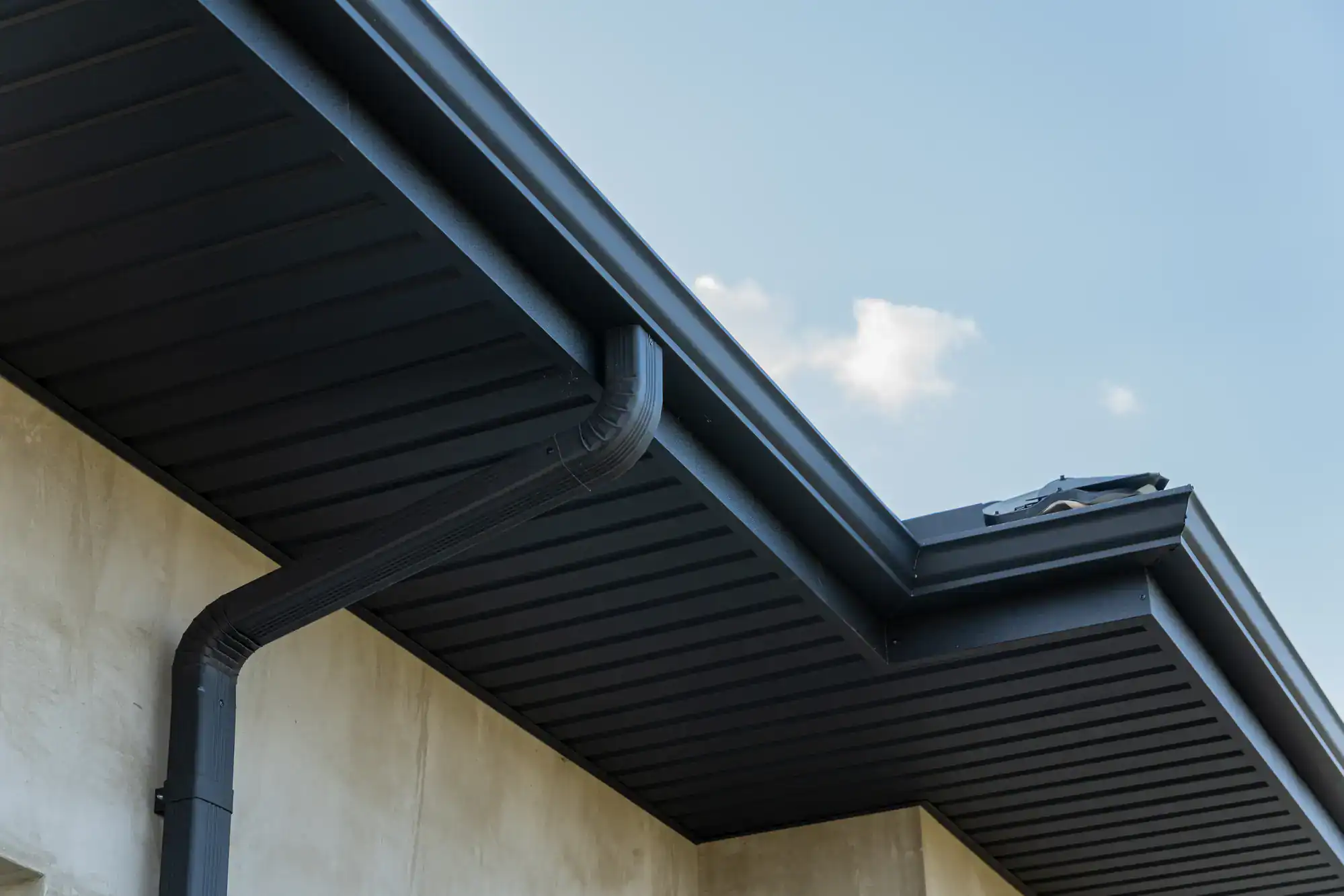 Close-up view of a modern house roof showing a black rain gutter system and downspout attached to the edge, with a light concrete wall and blue sky with a few clouds in the background.