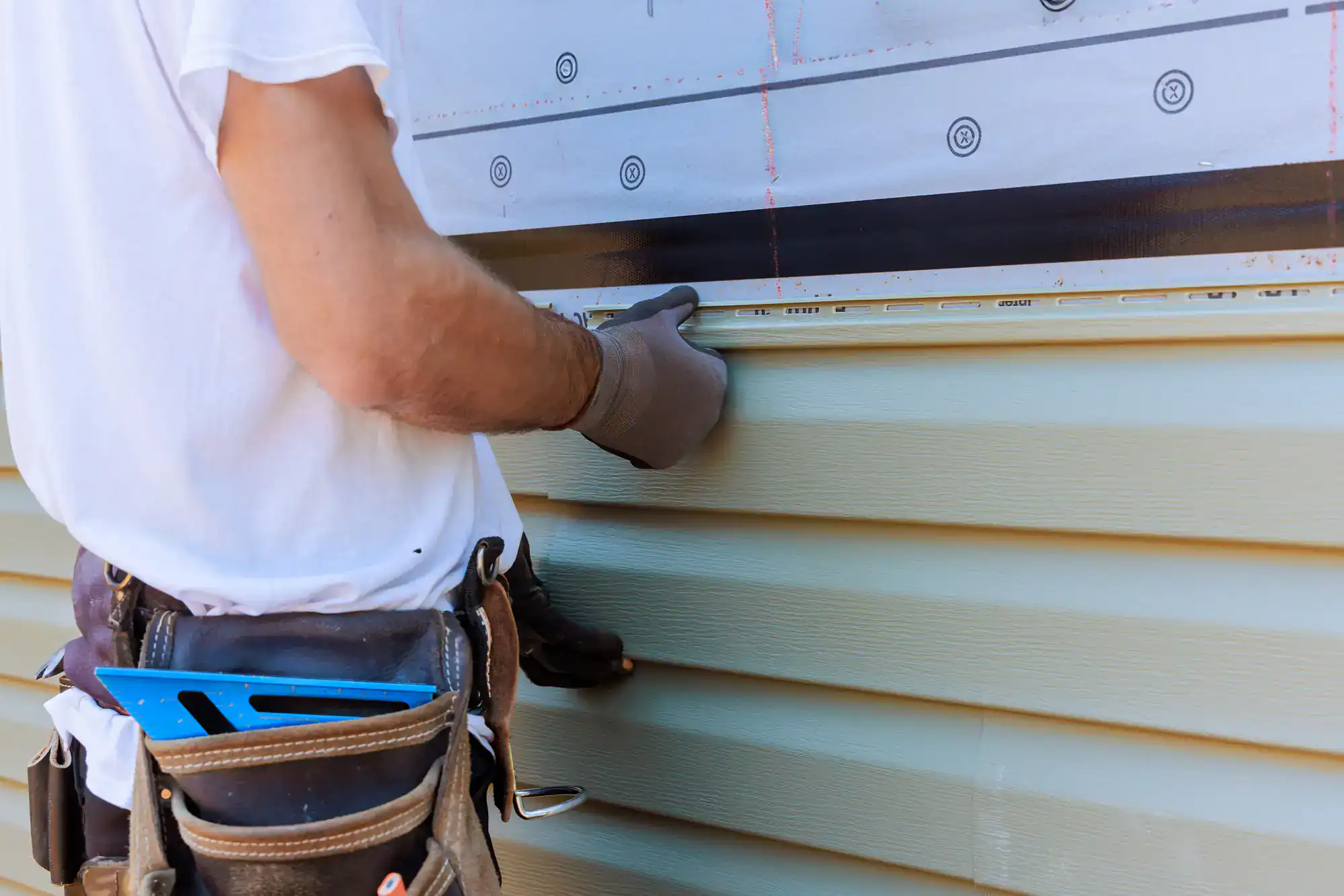 A person wearing gloves and a tool belt installs light green vinyl siding on a house, using a level to ensure proper alignment. Only their torso and arms are visible.