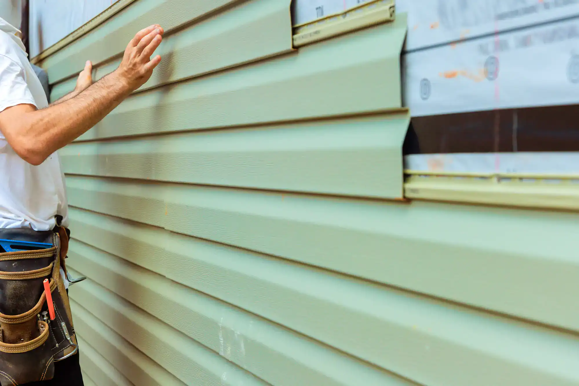 A person wearing a tool belt checks light green vinyl siding that is being installed on the exterior wall of a house, with one section unfinished and covered with protective material.