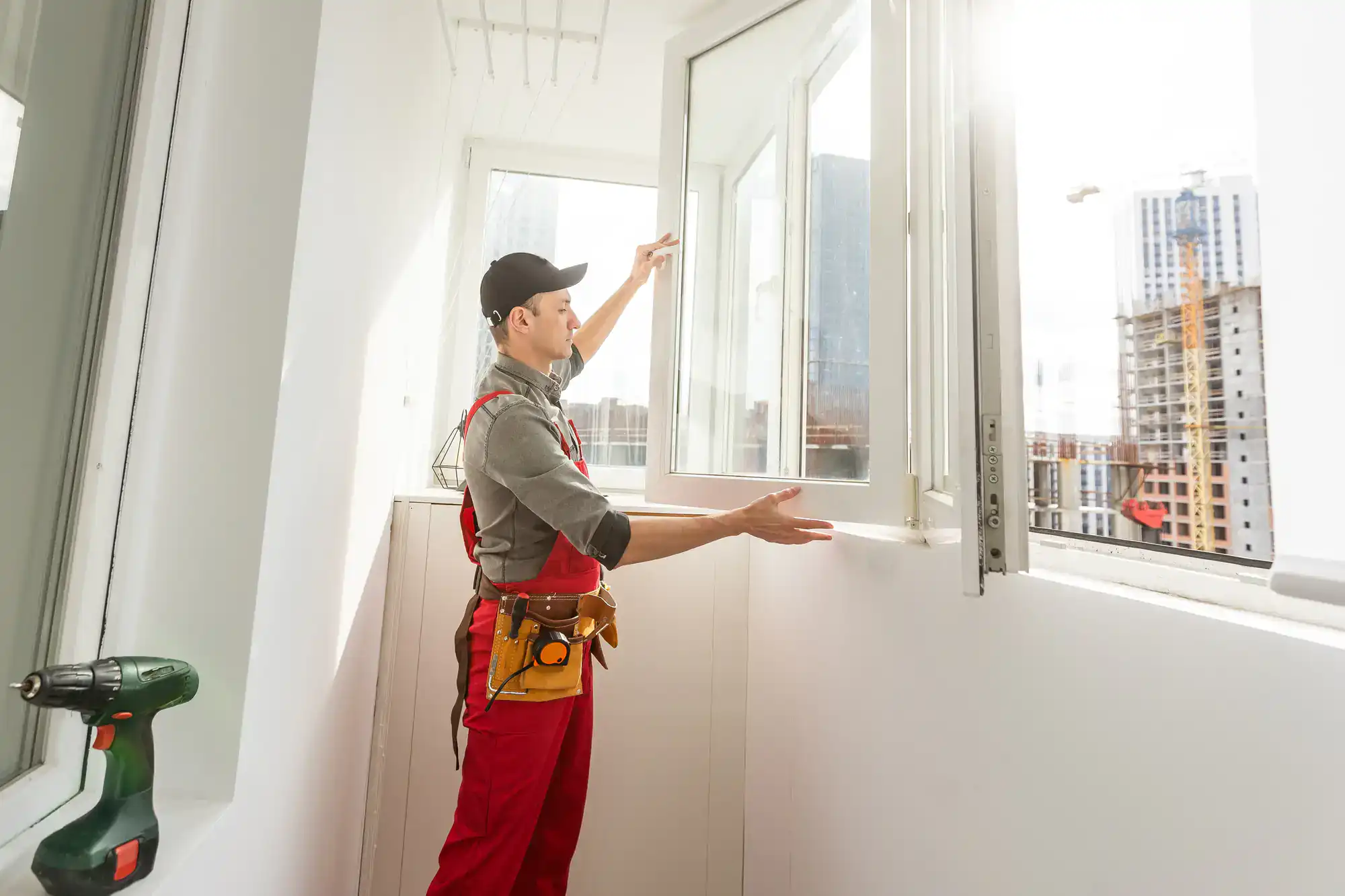 A worker in red overalls and a black cap installs a large window in a bright room, with a cityscape and construction cranes visible outside. A green power drill rests on a nearby surface.