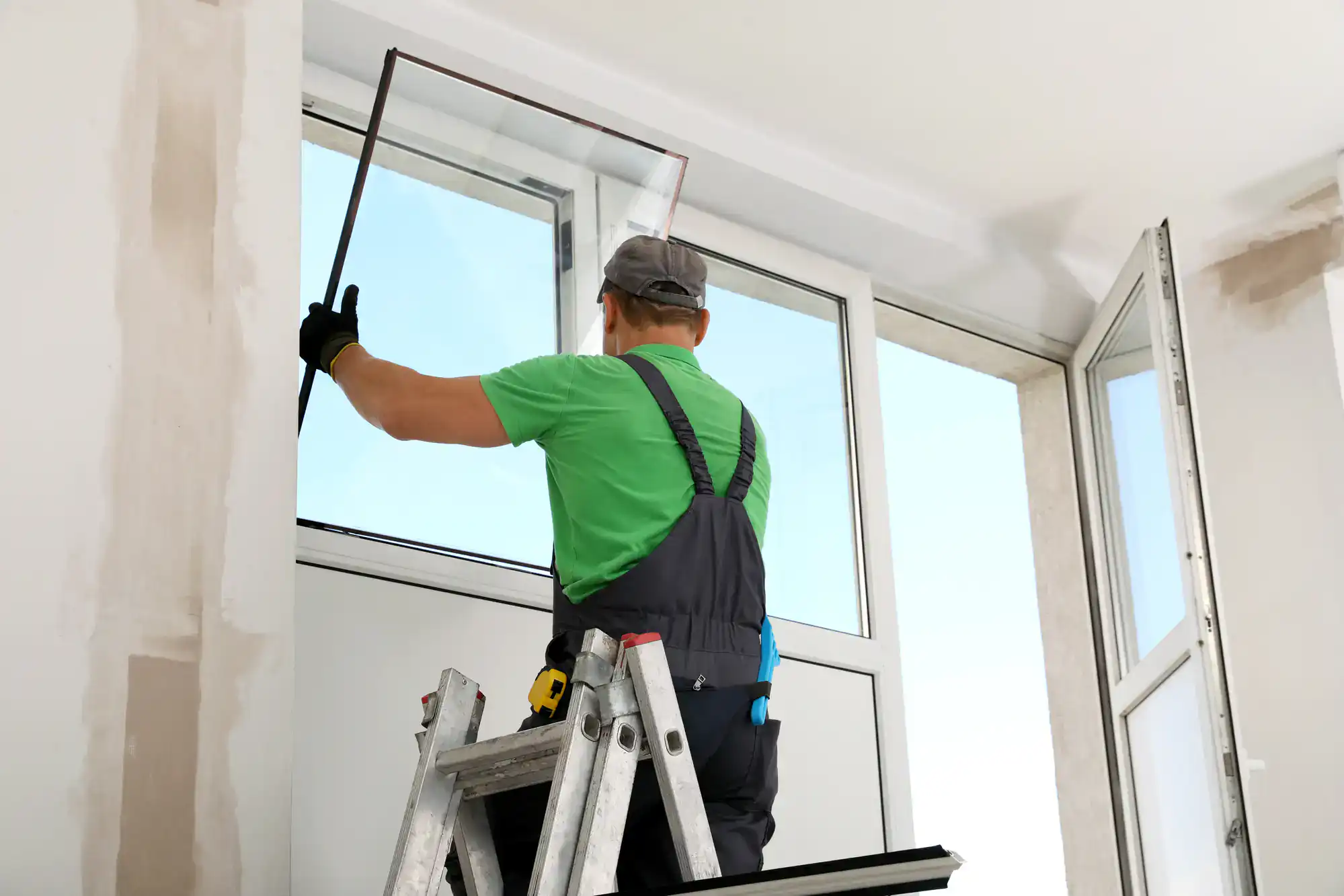 A worker in a green shirt and overalls stands on a ladder, installing or repairing a window screen in a bright indoor space with white walls.