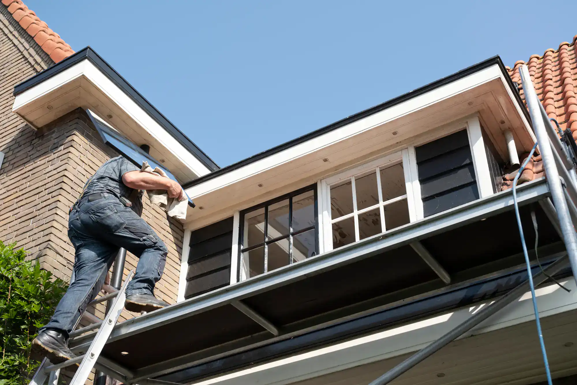 A worker standing on a ladder is repairing or installing siding and trim beneath the roof of a house, with scaffolding below and a clear blue sky above.