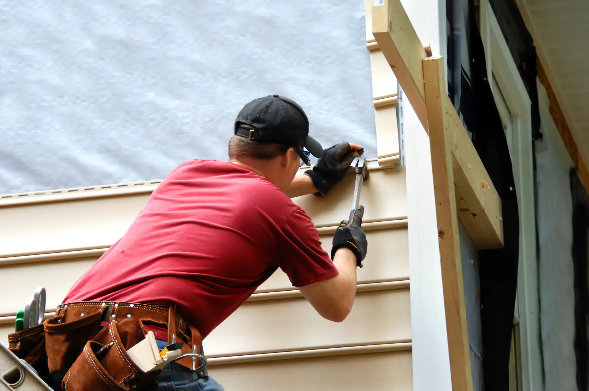 A construction worker wearing a red shirt and black cap installs beige vinyl siding on a house, using a hammer and nails. He has a tool belt and faces the wall, working on an exterior renovation.