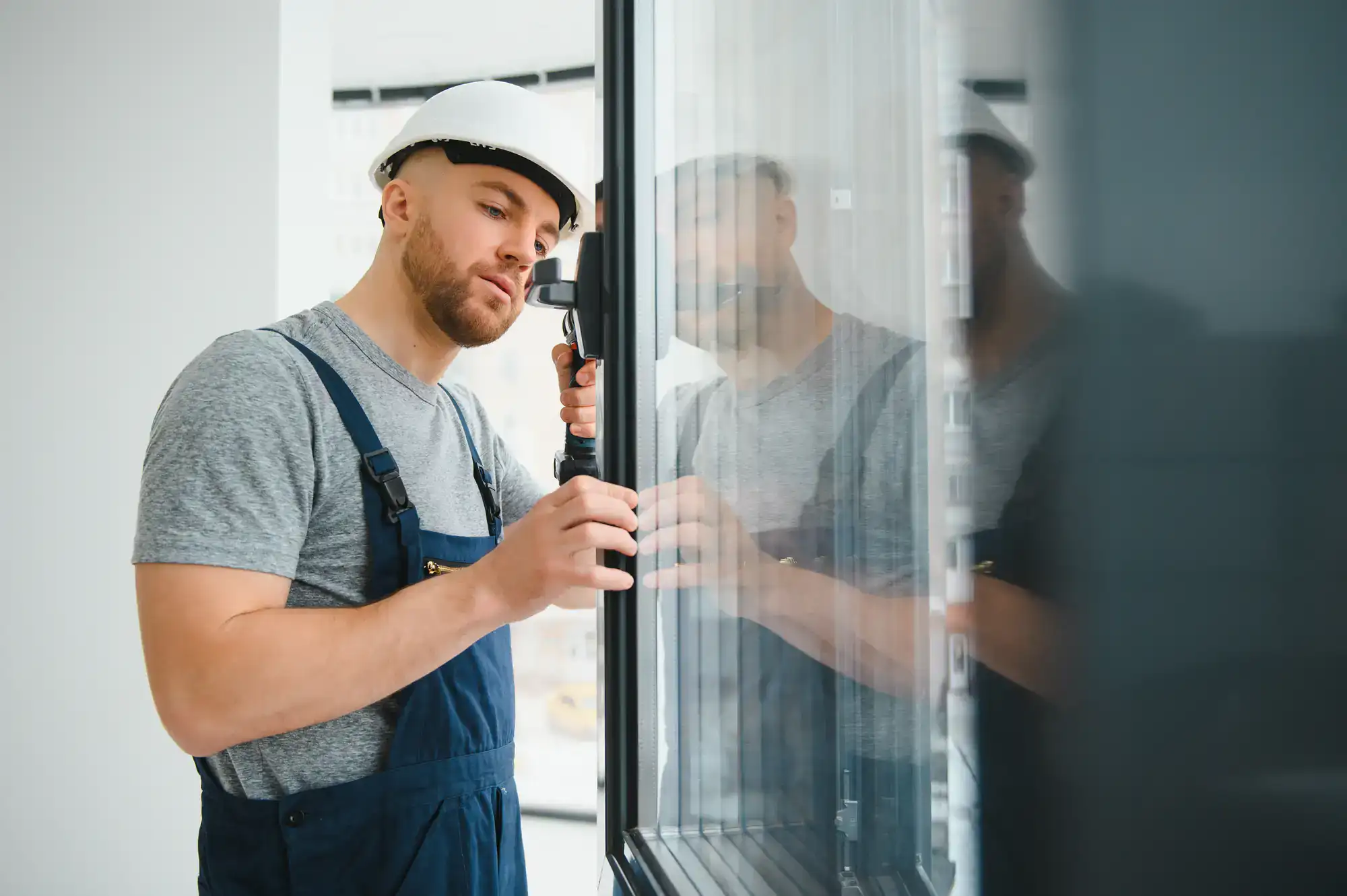 A construction worker in overalls and a hard hat inspects or installs a window, carefully adjusting the frame inside a modern building.