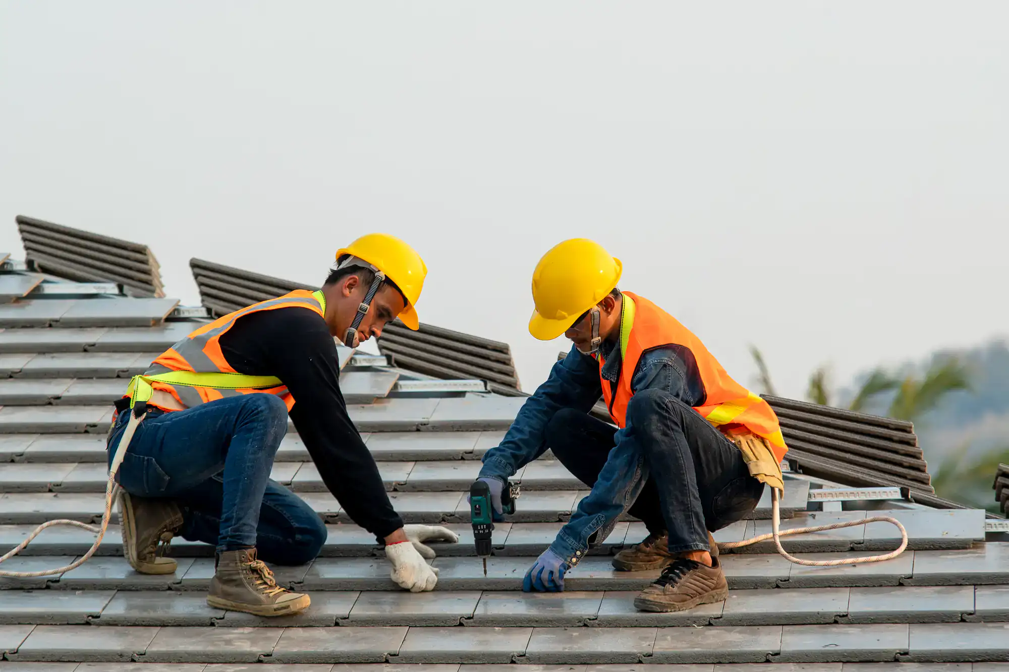 Two construction workers wearing yellow helmets and orange safety vests install roof tiles on a building, using tools and protective gear while crouched on the sloped roof.