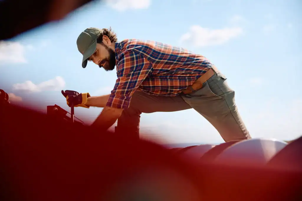 A man wearing a cap, plaid shirt, and gloves works outdoors, kneeling on a metal surface under a blue sky with clouds. Some red objects are blurred in the foreground.
