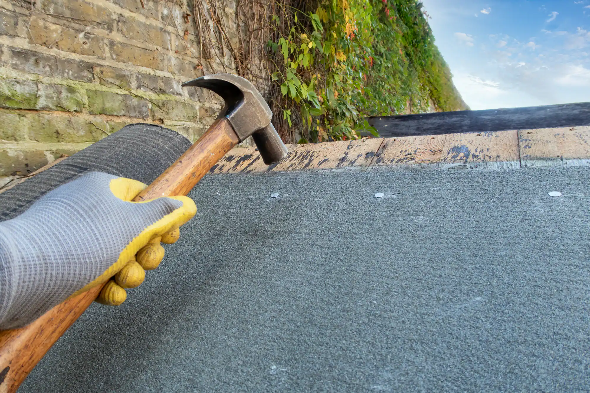 A gloved hand holds a hammer over a partially finished roof, preparing to secure roofing material. A brick wall with green ivy is in the background under a blue sky.