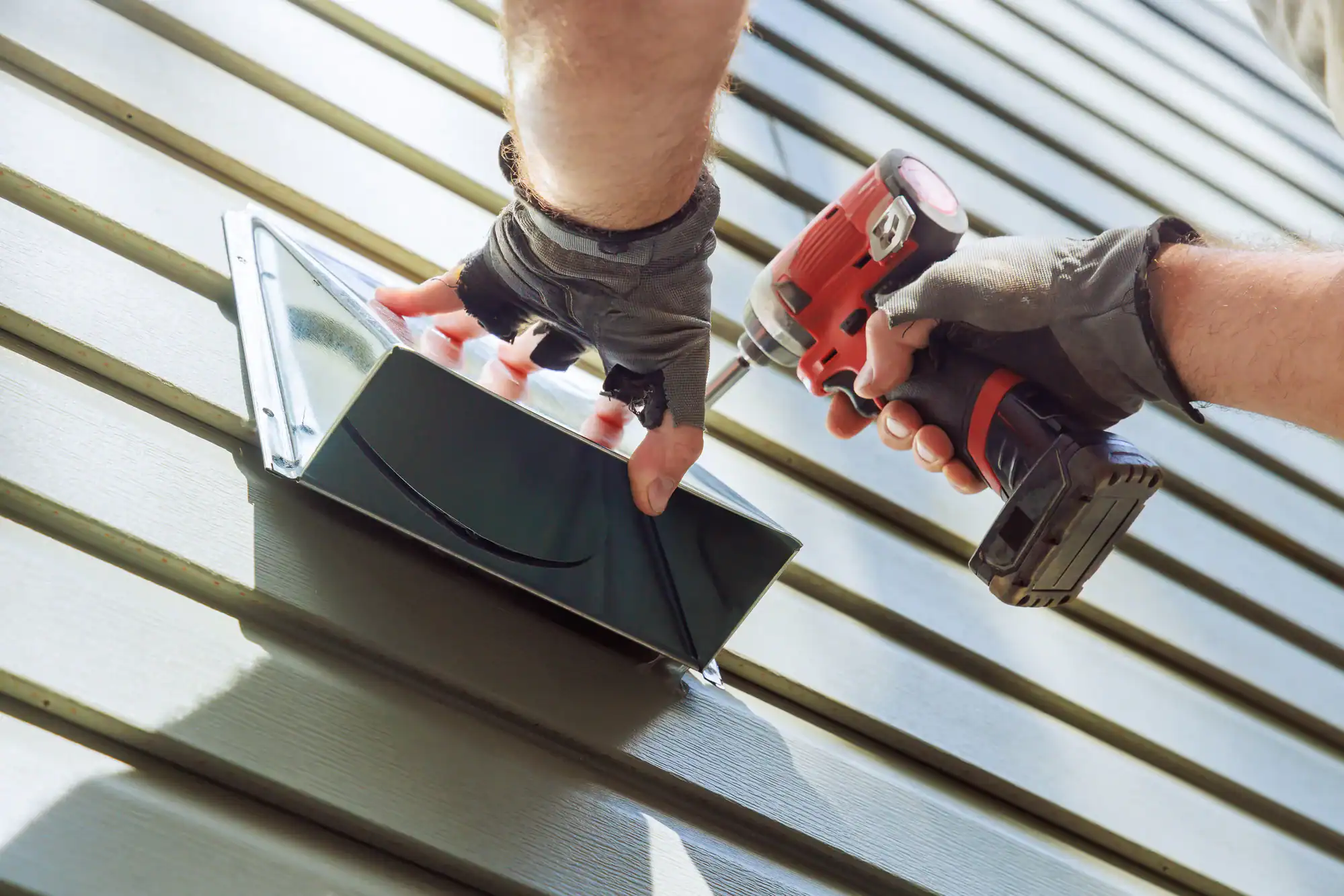 A person wearing gloves uses a cordless drill to install or repair a vent cover on the exterior wall of a house with horizontal siding.