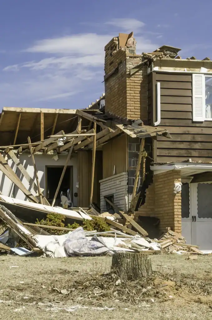 A partially collapsed house with exposed wooden beams and bricks, significant roof and wall damage, and debris scattered on the ground in front. The sky is clear and blue.