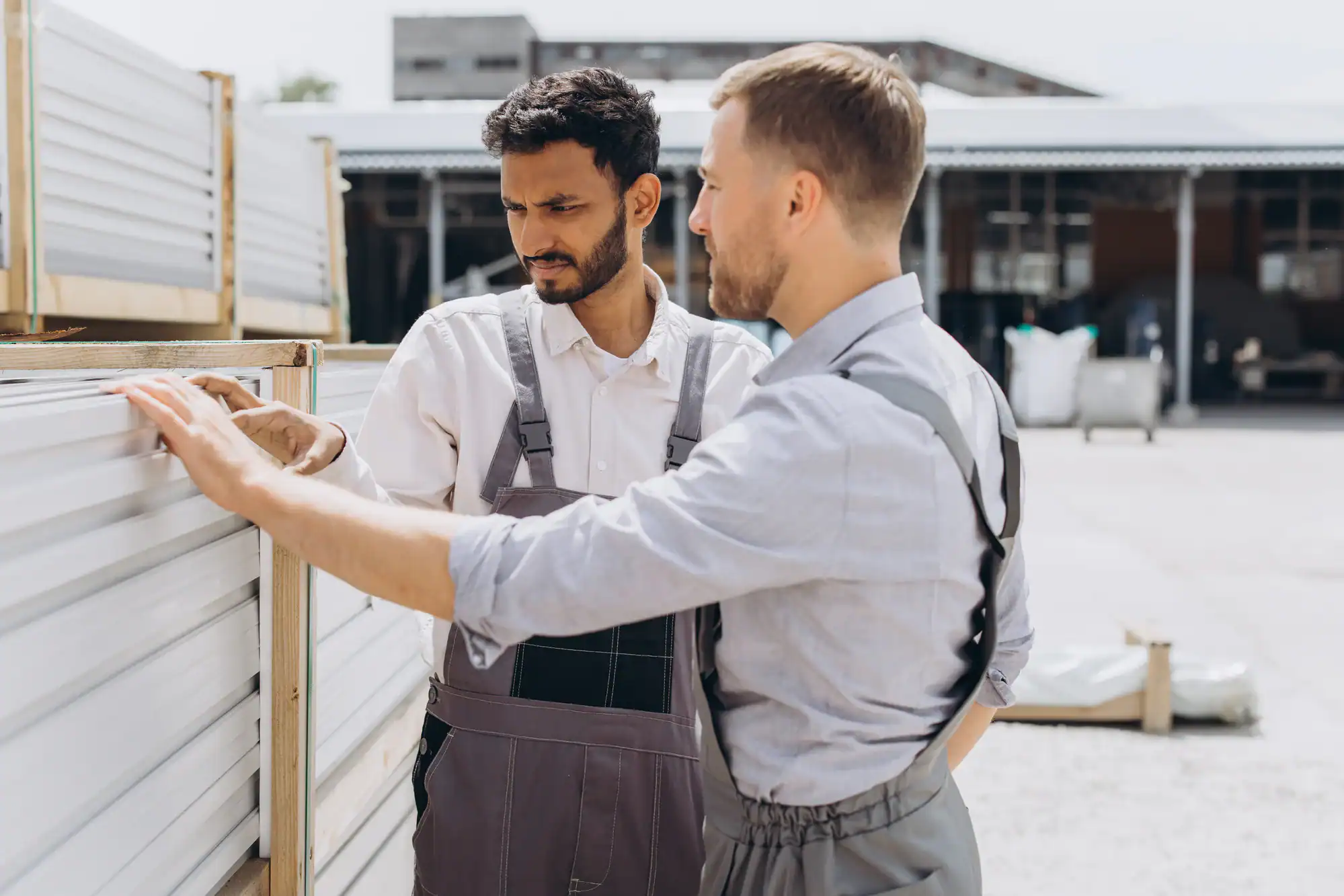 Two men wearing work overalls inspect and discuss a white metal panel outdoors at an industrial or construction site, with buildings and equipment visible in the background.