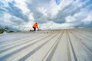 A worker in an orange shirt and yellow helmet kneels on a large metal rooftop under a cloudy sky, appearing to perform maintenance or repairs.