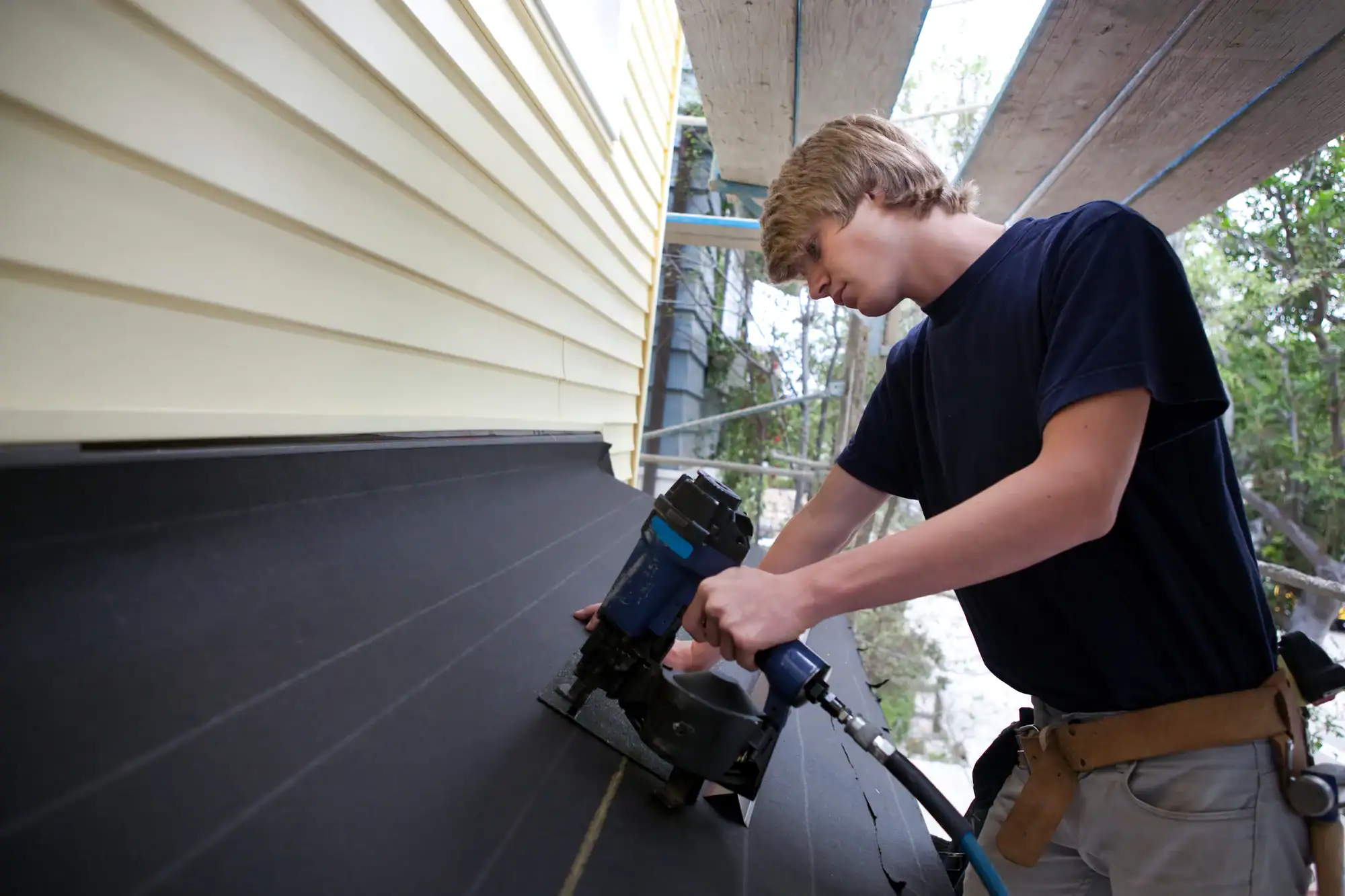 A person wearing a tool belt uses a nail gun to secure roofing material on a sloped roof next to a yellow house, with scaffolding and trees in the background.