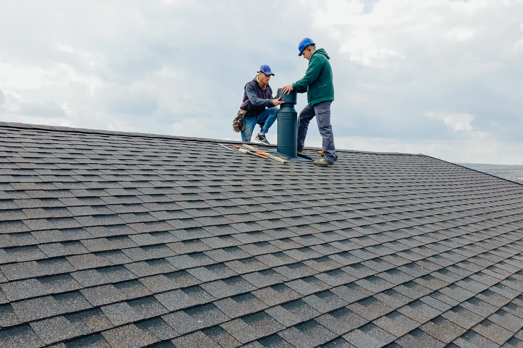 Two workers in blue caps are repairing or installing a vent on a shingled house roof. Tools are laid out beside them, and the sky is cloudy in the background.